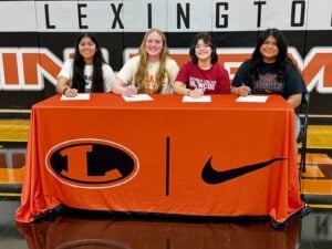 Four Lexington Minutemaids sitting at a table with orange and black decorations for their college athletic signing ceremony.